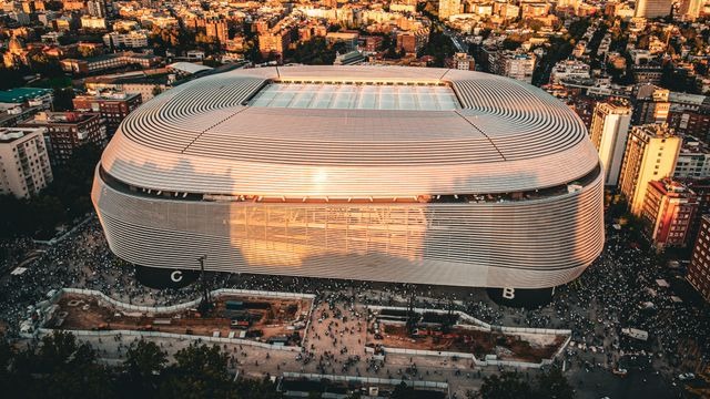 Moderan stadion sa zaobljenom metalnom fasadom okružen širim gradom i gomilom ljudi