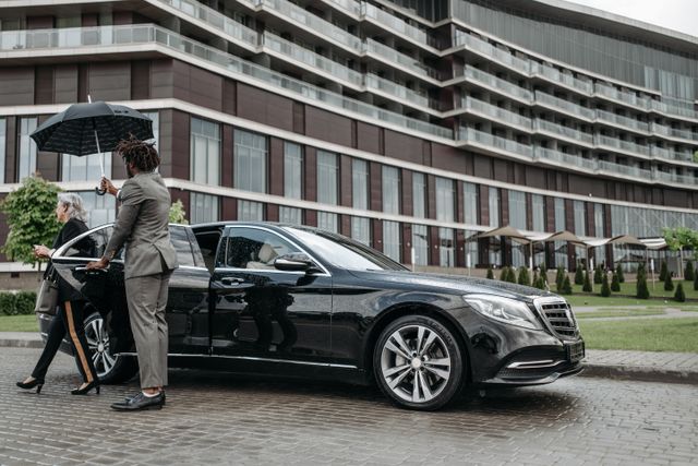 Muškarac drži kišobran nad ženom koja izlazi iz crnog automobila ispred moderne zgrade